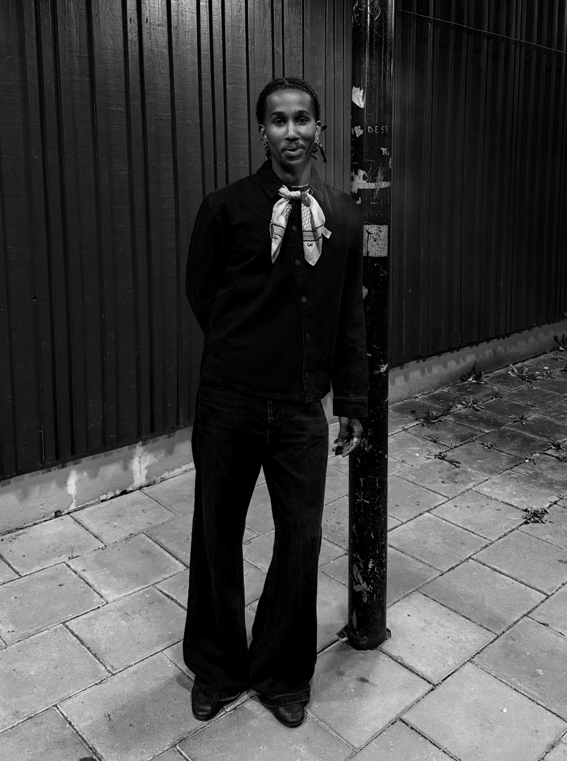 Nabil Nassor standing against a dark wooden fence and a lamp post, in black clothes with a light silk scarf, black and white photo
